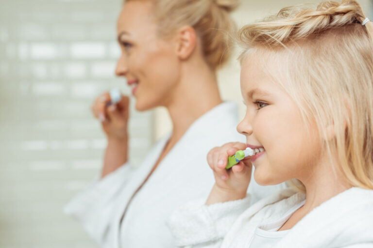 Side View Of Happy Mother And Daughter In Bathrobes Brushing Teeth Together Preventative dental image: brushing teeth